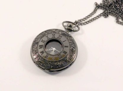 Gun metal black fob watch - closed - with neck chain. The cover is etched with a swirling pattern and it has roman numerals etched around a centre clear panel showing the watch underneath